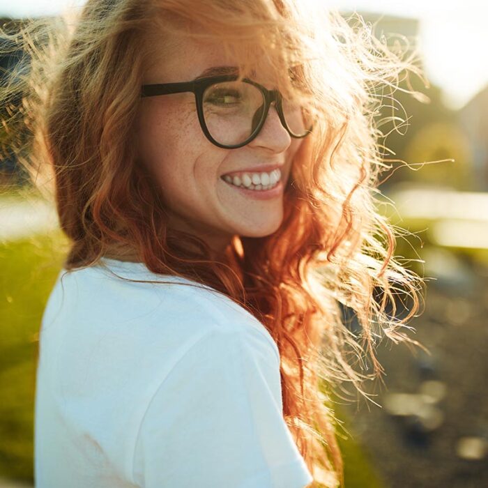 young smiling woman with glasses outdoors on a sunny day looking back over her shoulder at camera